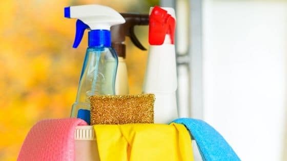 Plastic bucket filled with cleaning supplies including spray bottles, sponges, and gloves